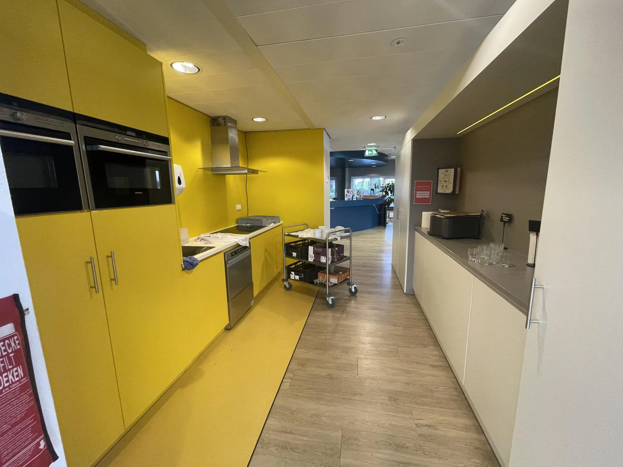 Moderne kantoorpantry met gele keukenwand, ingebouwde apparatuur en een serveerwagen in een gang aan de Jan Leentvaarlaan.